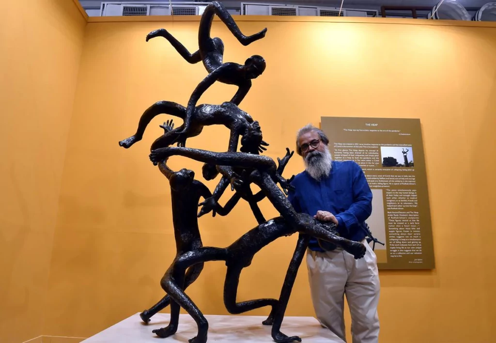 KS Radhakrishnan stands in front of orange background, next to one of his life-size sculptures. 